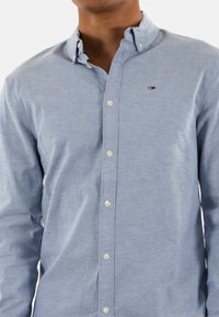 Camisa azul claro de botones con cuello extendido, con botones blancos y un pequeño logotipo bordado. Fabricada con un tejido texturizado.