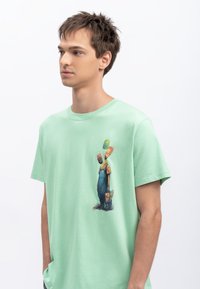 T-shirt en coton vert clair avec une coupe décontractée. Présente un design graphique de personnages en salopette sur le devant. Texture douce.