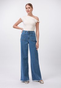 Bela top z enim ramenom in nabranim detajlom, v kombinaciji z visokotassisvimi širokimi denim hlačami v modri barvi, ki imajo sprednje žepe in neobdelan rob.