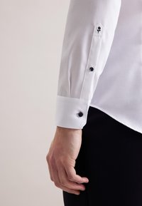 Camicia bianca con polsini a punta, caratterizzata da bottoni neri e un dettaglio ricamato a forma di rosa vicino al polsino. Tessuto liscio e vestibilità sagomata.