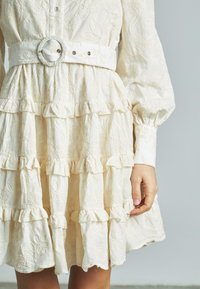 Weißes, gestuftes Kleid mit floraler Stickerei, Rüschensaum und langen Ärmeln. Kombiniert mit einem breiten Gürtel, der eine runde silberne Schnalle hat.