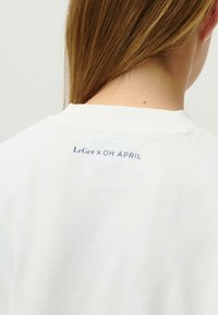 Balta medvilninė marškinėlių su apvaliu kaklu ir trumpomis rankovėmis, su mėlynu tekstu "LeGer x OH APRIL" nugaroje, šalia apykaklės.