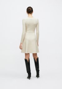 Κρεμ φόρεμα μήκους μέχρι το γόνατο με υφασμένο ανάγλυφο πλεκτό μοτίβο, εφαρμοστό μπούστο, μακριά μανίκια και φουσκωτή φούστα· συνδυασμένο με μαύρες μπότες μέχρι το γόνατο.