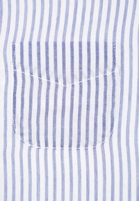 Witte stof met blauwe verticale strepen heeft een kleine rechthoekige borstzak. De stiksels zijn zichtbaar langs de rand van de zak.