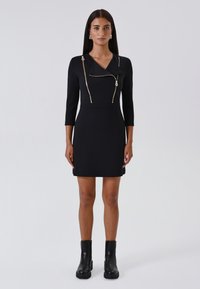 Schwarzes, tailliertes Kleid mit drei Viertelärmeln, das mit einem dekorativen goldfarbenen Reißverschluss und Kragenverzierungen versehen ist. Getragen mit schwarzen Stiefeletten.