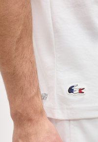 Κοντινό πλάνο στο μπράτσο ενός ατόμου δίπλα στο τελείωμα ενός λευκού πουκάμισου που φέρει ένα μικρό λογότυπο κροκόδειλου σε μπλε, λευκό και κόκκινο χρώμα.