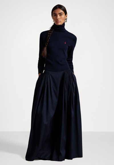 Sieviete ar gariem pinumiem, tērpusies tumši zila augsta kakla džemperī un pilnā, pleatotā tumši zilā garā svārkā, stāv ar rokām kabatās.