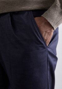 Marine bukser med en glat tekstur, der har en sidelomme og en brun hånd indsat, accentueret af en beige sweater ovenfor.