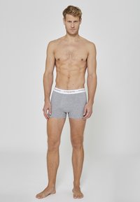Boxer briefs en coton gris avec une ceinture blanche ornée de texte noir. Le modèle est pieds nus, debout devant un fond uni.