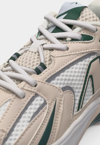 Beež ja valge võrksest spordijalats roheliste aktsentidega, valged paelad on roheliste triibuliste aukude kaudu nööritud ning on nähtav polsterdatud keel.