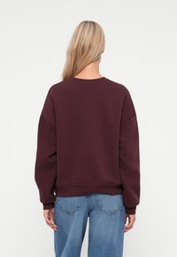 Sweatshirt cor castanha com um corte descontraído, decote redondo e ombros caídos. Textura suave com punhos e bainha canelados. Usado com jeans de cor azul claro.