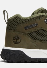 Zielone zamszowe i siateczkowe buty trekkingowe z czarną teksturowaną podeszwą, z naszywką logo i metalowym oznakowaniem po boku.
