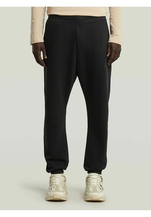 Persona che indossa pantaloni della tuta neri con polsini elastici e sneakers beige, in piedi davanti a uno sfondo chiaro e uniforme.