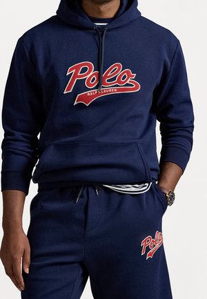 Un homme porte un sweat à capuche bleu marine et un pantalon de survêtement avec des logos rouges « Polo Ralph Lauren », une chemise rayée qui dépasse, ainsi qu'une montre-bracelet à la main gauche.