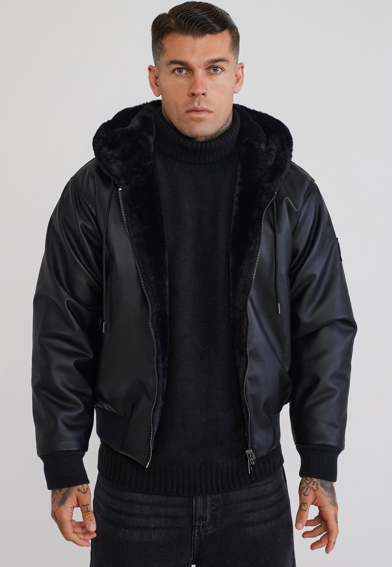 Homme portant une veste en similicuir noire avec capuche bordée de fourrure, un pull à col roulé noir et un jean foncé, debout devant un fond uni.