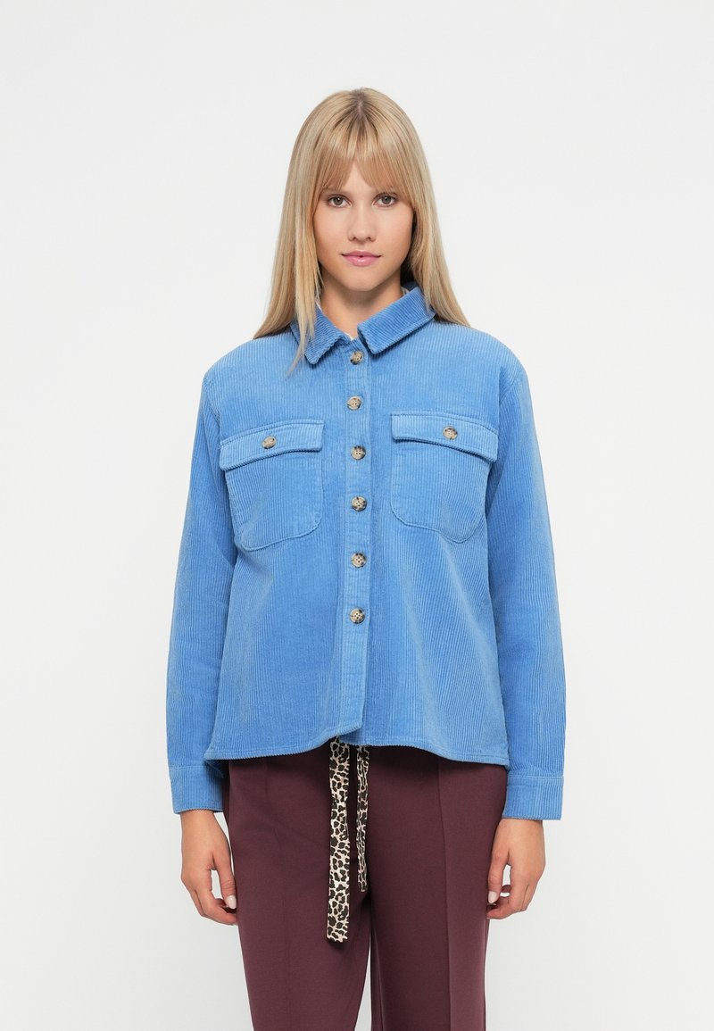 Ανοιχτό μπλε καπιτονέ πουκάμισο με κουμπιά, διαθέτει δύο μπροστινές τσέπες και μεταλλικά κουμπιά. Έχει γιακά και ελαφρώς κομμένη αθλητική γραμμή στο στρίφωμα.