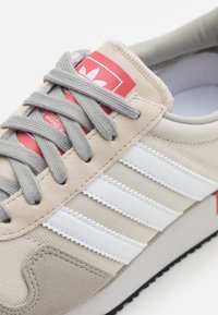 Beige Adidas-sneakers med grå snören, vita tre ränder och en röd logotag på tungan, som visar närbilder av materialen.