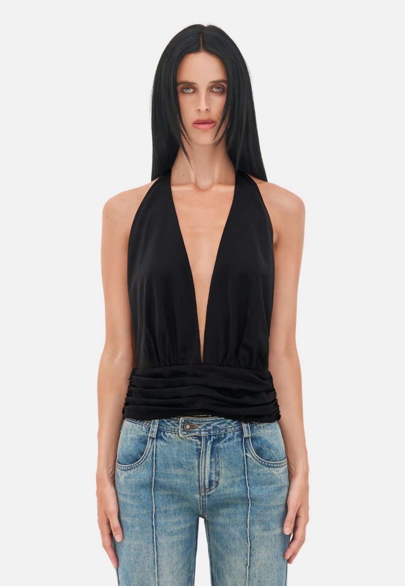 Woman with long black hair wearing a sleeveless black halter top with deep V-neck and blue high-waisted jeans standing against white background.