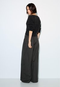 Vrouw met kort donker haar, gekleed in een zwarte, pluizige trui en wijde donkergrijze broek, staand tegen een witte muur, terwijl ze achterom kijkt.