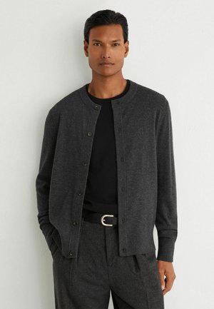 Uomo che indossa un cardigan grigio scuro abbottonato sopra una camicia nera con pantaloni grigio scuro abbinati e cintura nera, in piedi contro un muro bianco.