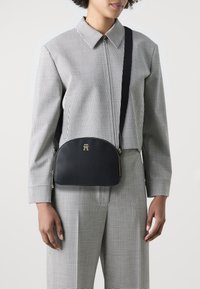 Borsa a tracolla in pelle nera con una forma curva, hardware dorato e un logo sul davanti, abbinata a una camicia a quadri e pantaloni.