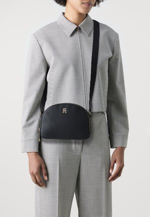 Borsa a tracolla in pelle nera con una forma curva, hardware dorato e un logo sul davanti, abbinata a una camicia a quadri e pantaloni.