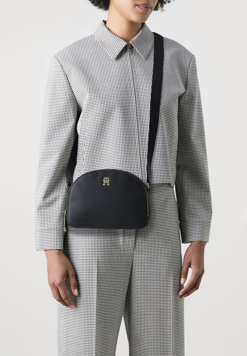 Borsa a tracolla in pelle nera con una forma curva, hardware dorato e un logo sul davanti, abbinata a una camicia a quadri e pantaloni.