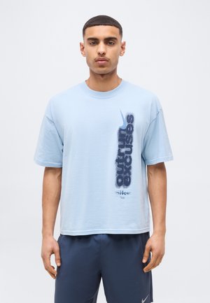 Fiatal férfi világoskék Nike pólóban, amelyen függőleges "outrun excuses" felirat és egy Nike logó található, sötétkék rövidnadrággal párosítva.