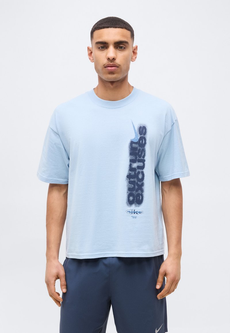 Νεαρός άνδρας που φοράει ανοιχτό μπλε μπλουζάκι Nike με κάθετο κείμενο "outrun excuses" και λογότυπο swoosh, συνδυασμένο με σκούρα μπλε σορτς.