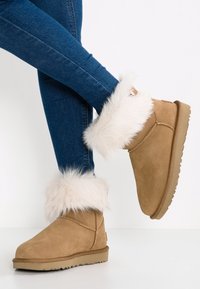 Bottines en daim marron clair avec bordure en fausse fourrure blanche, bout arrondi, semelle plate et coutures contrastantes. Portées avec un jean skinny bleu foncé.