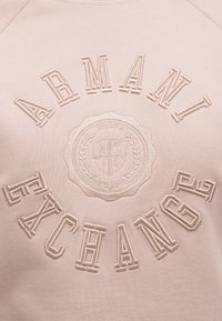 Το ARMANI EXCHANGE κεντημένο με μεγάλα γράμματα γύρω από ένα κυκλικό έμβλημα με το MILANO AX 91 EST. 1991 σε μπεζ ύφασμα.