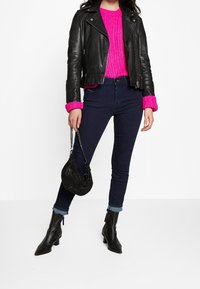 Chaqueta de cuero negra de motorista, suéter de punto rosa brillante con puños acanalados, jeans ajustados oscuros, botas de tobillo negras y un bolso negro decorado con cuentas.