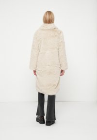 Garšs, kažokām līdzīgs bēša mētelis ar augstu apkakli, izgatavots no mīksta materiāla. Mētelis ir brīvi pieguļošs un sasniedz vidussilti, apvienots ar izsistām pelēkām biksēm.