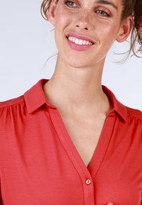 Red blouse with a collared neckline and button detail. Soft, textured fabric with subtle gathers at the shoulders.