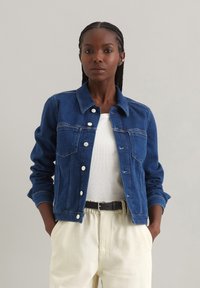 Blauwe spijkerjack met een kraag, zilveren knopen, twee voorkeetzakken en lichte slijtage, gedragen over een witte blouse en lichte broek.