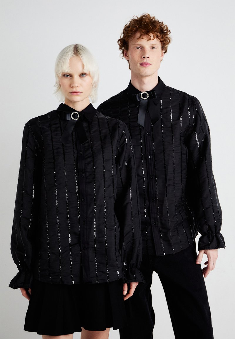 Las camisas negras abotonadas presentan una tela texturizada con rayas verticales de lentejuelas, mangas largas y acentos de pajarita en el cuello.