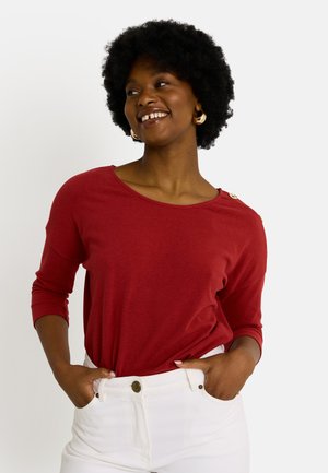 Haut rouge à manches 3/4 avec un col large, texture lisse et un détail de bouton décoratif à l'épaule, assorti à un jean blanc.