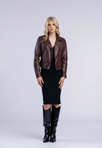 Blonde vrouw in een bruine leren jack, een zwart geribbeld midi-jurk en zwarte kniehoge laarzen, staand tegen een effen achtergrond.