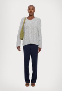 Gray knitted sweater with a V-neck, textured cable design, paired with navy trousers and brown shoes. Green crossbody bag adds color.