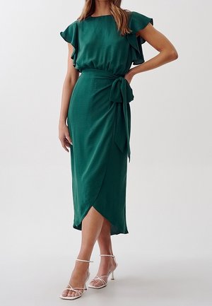 Femme portant une robe midi verte avec des manches volantées et une ceinture nouée à la taille, associée à des sandales blanches à talons et à brides.