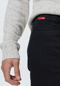 Main à côté de la hanche, portant un jean noir avec une étiquette rouge et un pull beige tacheté dont les manches sont légèrement retroussées.