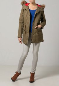 Olivgrön parka-jacka med en röd fodrad huva, med pälskant, framfickor och justerbara snoddar. Kombineras med ljusgrå jeans och bruna ankelskålar.