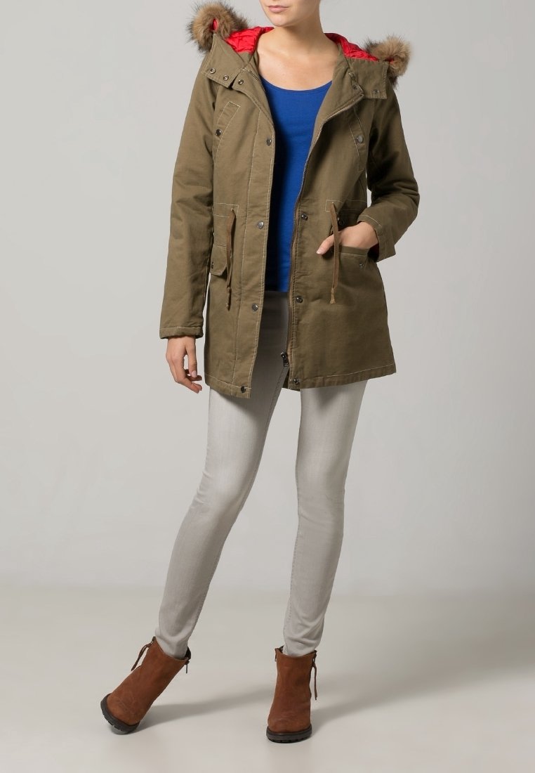 Olivgrön parka-jacka med en röd fodrad huva, med pälskant, framfickor och justerbara snoddar. Kombineras med ljusgrå jeans och bruna ankelskålar.