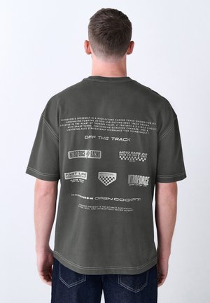 Mann trägt ein dunkelgrünes T-Shirt mit Rennsport-Thema, mehreren weißen Logos und Text auf dem Rücken, steht vor einem schlichten Hintergrund.