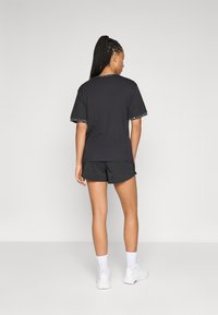 Kvinna med flätat hår står vänd bort med en svart t-shirt med mönstrad kant, svarta shorts, vita strumpor och vita sneakers.
