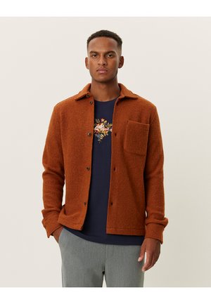 Rust-colored wool shirt jacket with a button front, chest pocket, and a textured finish, worn over a navy shirt with floral design.