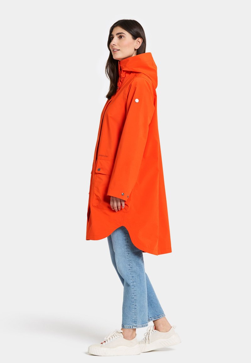 Orange wasserdichte Jacke mit Kapuze, im lockeren Schnitt, seitlichen Taschen und Druckknöpfen. Kombiniert mit hellblauen Jeans und weißen Sneakern.