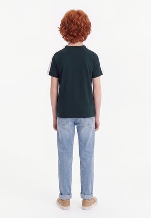 Bambino con capelli rossi ricci in piedi di spalle, indossa una maglietta verde scuro, jeans azzurri e sneakers beige.