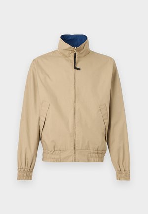 Veste beige légère avec col montant, fermeture éclair à l'avant, deux poches latérales, poignets élastiques et taille élastique, doublée de tissu bleu à l'intérieur du col.