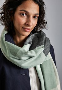Écharpe à carreaux verts et gris en mélange de laine douce, avec une finition texturée. Portée de manière décontractée sur une veste sombre et un pull léger.